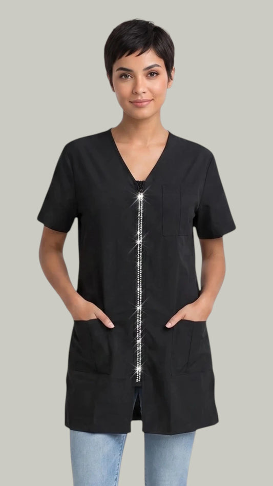 Front view of a short-haired woman posing with hands in pockets modeling the Ladybird Line 650 Rhinestone Zipper Water & Bleach Resistant Grooming & Styling Smock, featuring a short-sleeve black grooming jacket with a sparkling rhinestone zipper front, V-neckline, and deep front pockets, worn over light blue jeans against a light grey background.