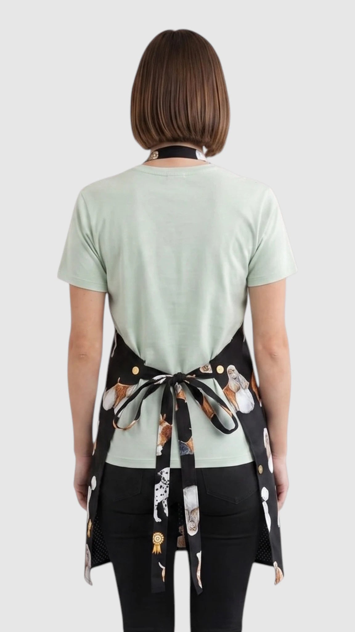 Back view of a short-haired woman modeling the Ladybird Line 655 Dog Groups Print Waterproof & Bleach Resistant Grooming Apron, showing the back tie belt closure tied at the waist over a mint green t-shirt and black pants against a light grey background.