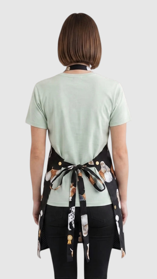 Back view of a short-haired woman modeling the Ladybird Line 655 Dog Groups Print Waterproof & Bleach Resistant Grooming Apron, showing the back tie belt closure tied at the waist over a mint green t-shirt and black pants against a light grey background.