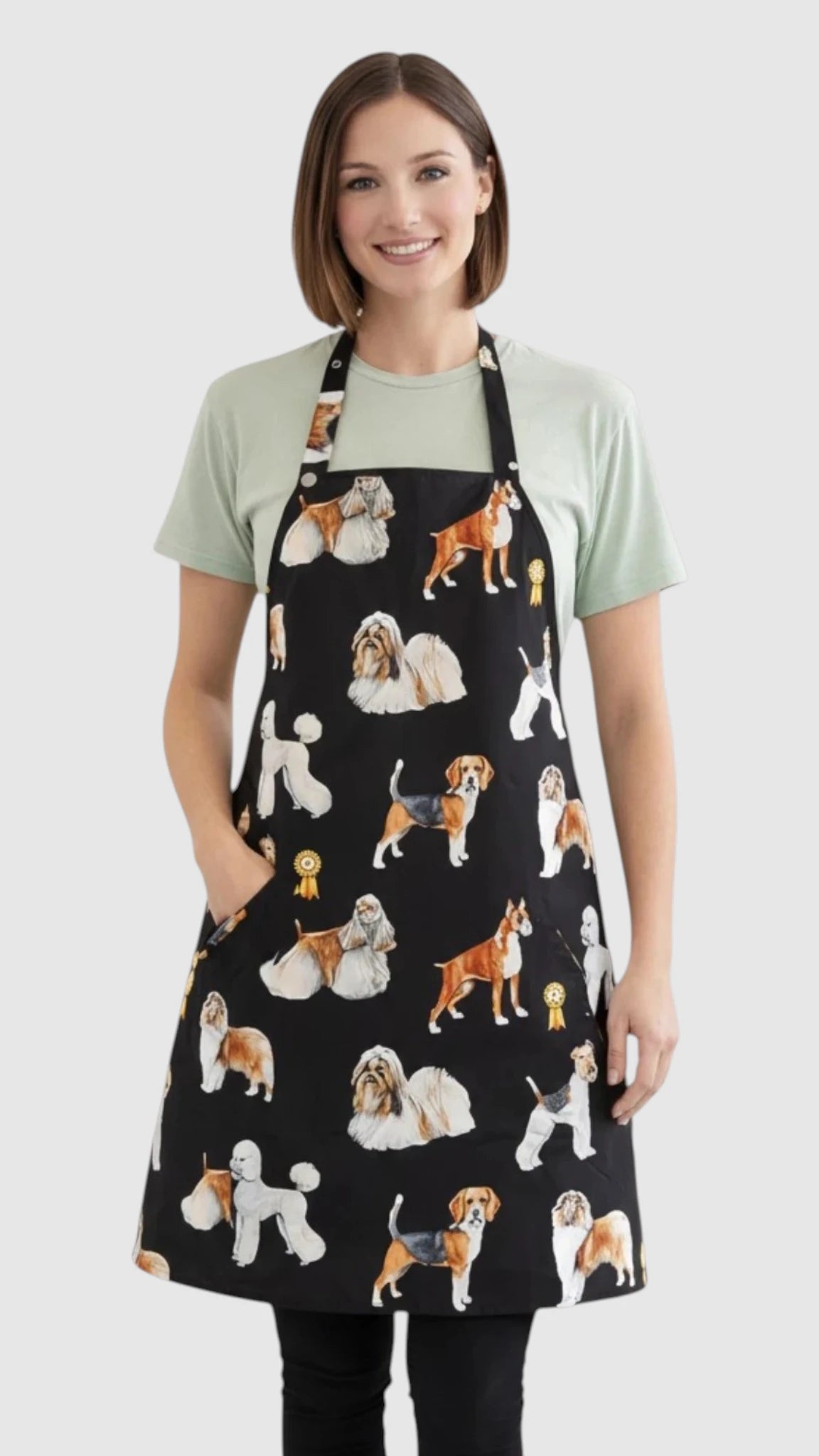 Front view of a smiling blonde woman posing with hand on hip modeling the Ladybird Line 655 Dog Groups Print Waterproof & Bleach Resistant Grooming Apron, featuring a full-coverage black apron with colorful portrait-style dog breed illustrations, adjustable neck strap, back tie belt, and deep front pockets, worn over black leggings against a light grey background.