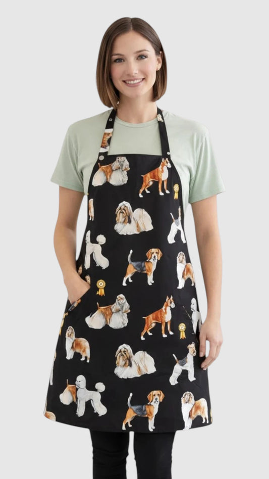 Front view of a smiling blonde woman posing with hand on hip modeling the Ladybird Line 655 Dog Groups Print Waterproof & Bleach Resistant Grooming Apron, featuring a full-coverage black apron with colorful portrait-style dog breed illustrations, adjustable neck strap, back tie belt, and deep front pockets, worn over black leggings against a light grey background.