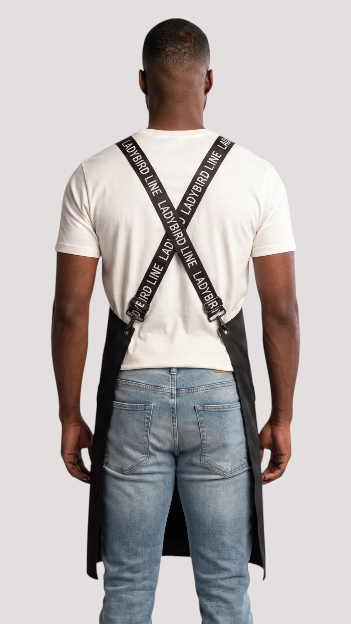 Back view of a man modeling the Ladybird Line 667 Waterproof Black Signature Strap Crossover Apron, showing the crisscross black signature straps printed with white 'LADYBIRD LINE' text over a white t-shirt and blue jeans against a solid off-white background.