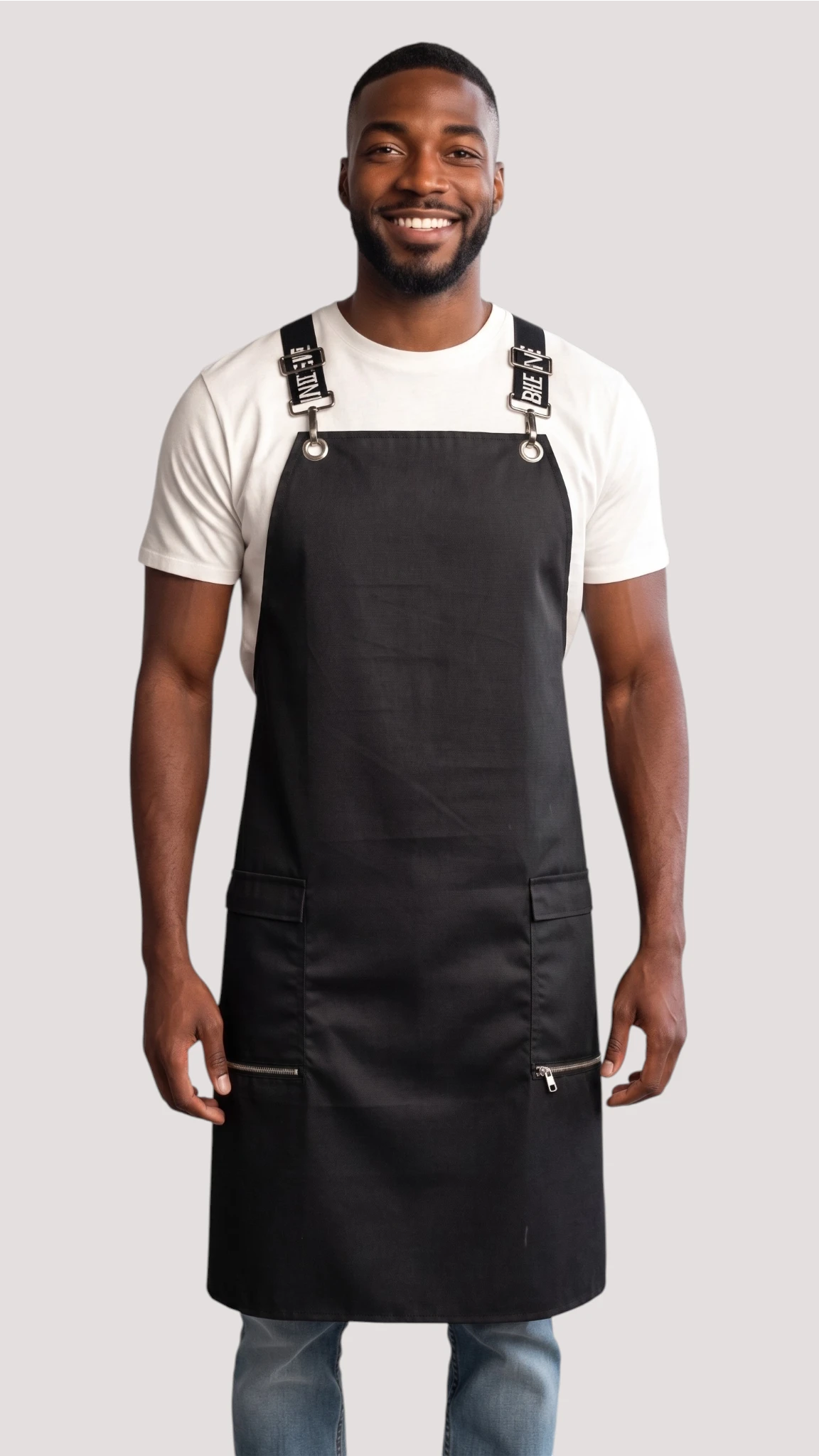 Front view of a man smiling while modeling the Ladybird Line 667 Waterproof Black Signature Strap Crossover Apron, featuring a solid black apron with black signature crossover straps with white text, zippered lapel pockets, worn over a white t-shirt and blue jeans against a solid off-white background.