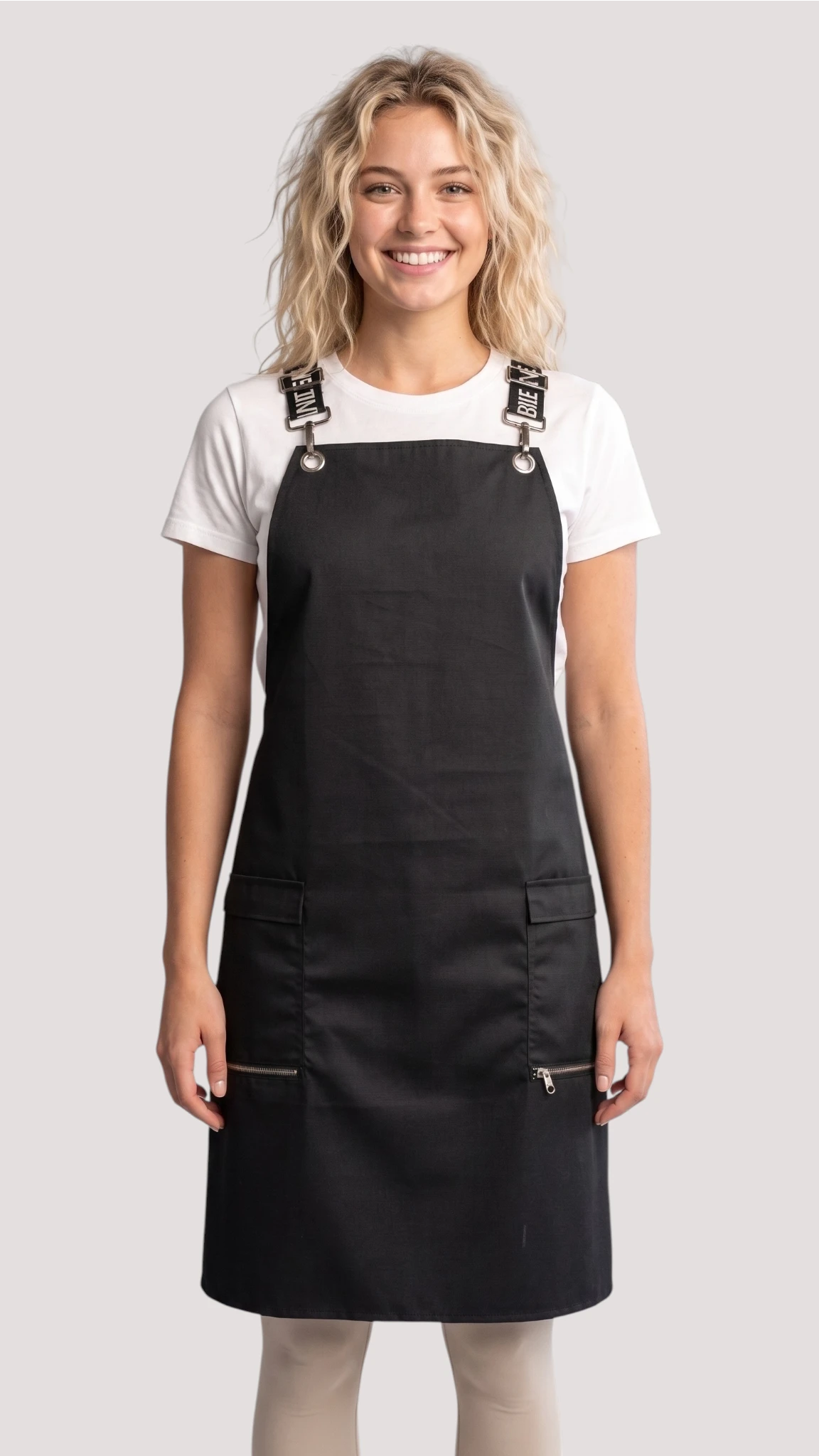 Front view of a smiling blonde woman modeling the Ladybird Line 667 Waterproof Black Signature Strap Crossover Apron, featuring a solid black apron with black signature crossover straps with white text, zippered lapel pockets, worn over a white t-shirt and light beige pants against a solid off-white background.
