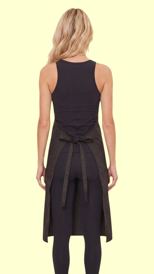 Back view of a blonde woman modeling the Ladybird Line 682 Bathers Long Black Waterproof Bathing Apron, showing the back tie closure cinched at the waist, worn over a black sleeveless top and black leggings against a light beige background.