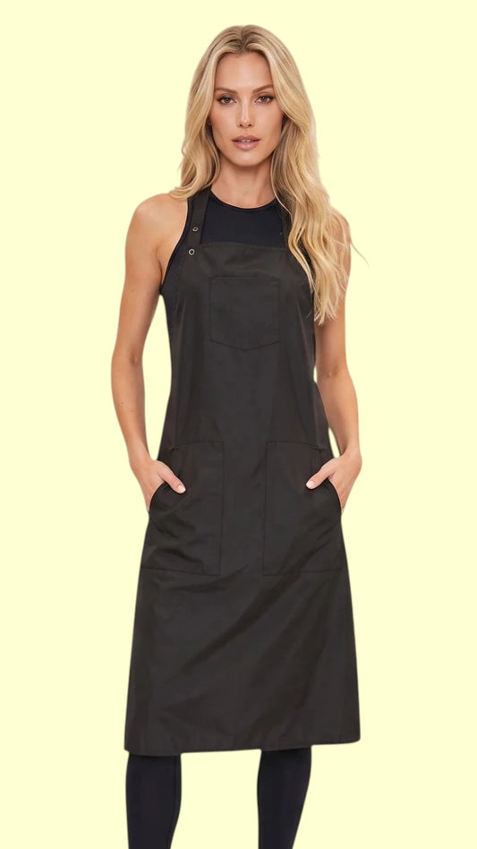 Front view of a blonde woman with hands in pockets modeling the Ladybird Line 682 Bathers Long Black Waterproof Bathing Apron, featuring a long black apron with deep side pockets, worn over a black top and black leggings against a light beige background.