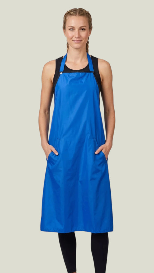 Front view of a blonde woman with braided hair smiling while modeling the Ladybird Line 680 Bathers Long Royal Blue Waterproof Bathing Apron, featuring a long royal blue apron with deep side pockets, worn over a black tank top and black leggings against a light beige background.
