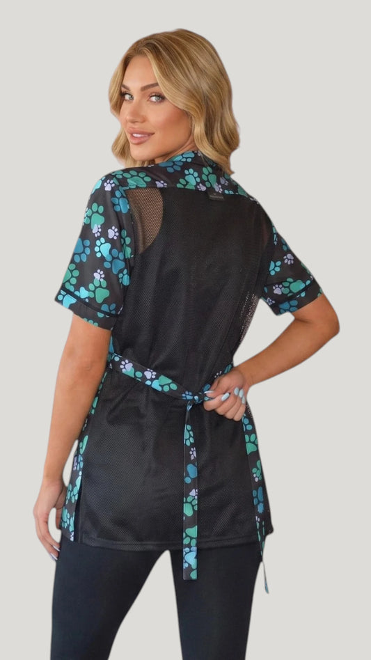 Back view of a blonde woman looking over her shoulder while modeling the Ladybird Line 701 Blue Paw Print Waterproof & Bleach Resistant Mesh Back Grooming Smock, showing the black mesh back panel, blue, teal, and purple paw print shoulder yoke, short sleeves, and back tie belt, worn over black pants against a light grey background.