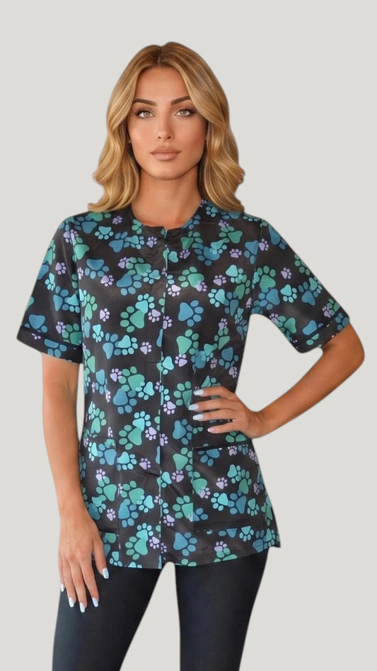 Front view of a blonde woman posing with hand on hip modeling the Ladybird Line 701 Blue Paw Print Waterproof & Bleach Resistant Mesh Back Grooming Smock, featuring a short-sleeve black grooming smock with a blue, teal, and purple paw print pattern, covered front zipper, and deep front pockets, worn over black pants against a light grey background.