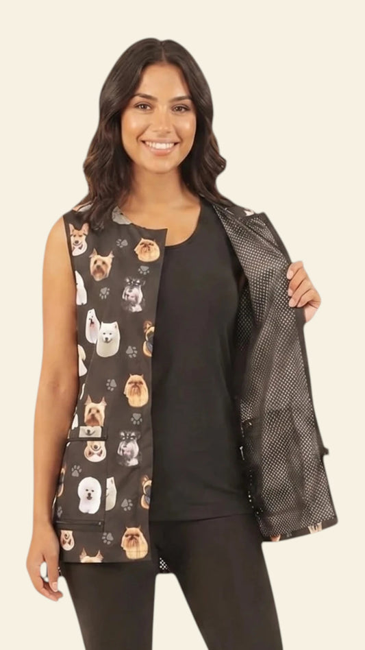 Front view of a smiling dark-haired woman holding open the front panel of the Ladybird Line 706 Dog Breeds Print Waterproof & Bleach Resistant Mesh Back Grooming Vest, showing the black mesh interior lining, colorful dog breed head print, covered front zipper, and deep flap pockets, worn over a black top and black pants against a solid light beige background.
