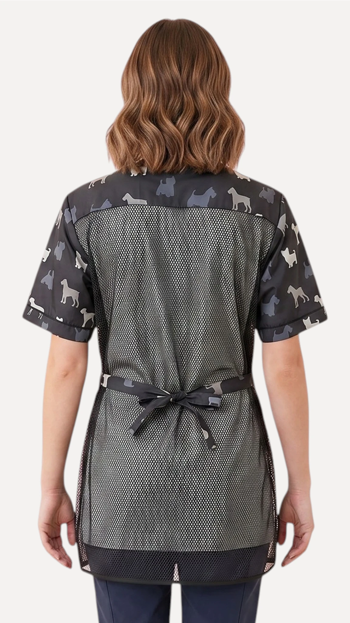Back view of a dark-haired woman modeling the Ladybird Line 709 Grey Terrier Unisex Waterproof Grooming Smock, featuring a full mesh back panel for ventilation, terrier silhouette print along the shoulders, sleeves, and tie belt, worn over dark pants against a solid white background.