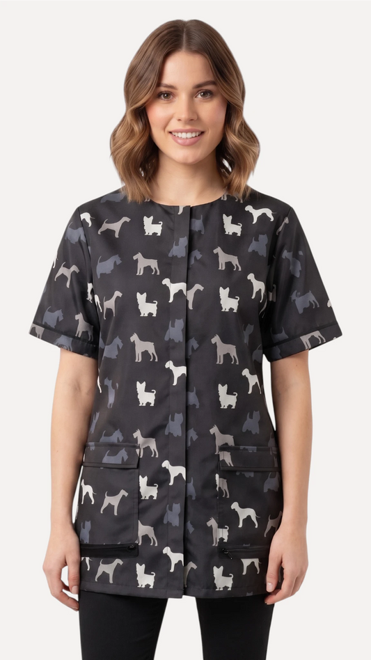 Front view of a dark-haired woman smiling while modeling the Ladybird Line 709 Grey Terrier Unisex Waterproof Grooming Smock, featuring an all-over grey, black, and white dog silhouette print with covered zipper lapels and front pockets, worn over black pants against a solid light grey background.