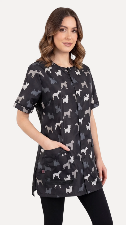 Side view of a dark-haired woman smiling while modeling the Ladybird Line 709 Grey Terrier Unisex Waterproof Grooming Smock, featuring an all-over grey, black, and white dog silhouette print with hands in side pockets, worn over black leggings against a solid light grey background.