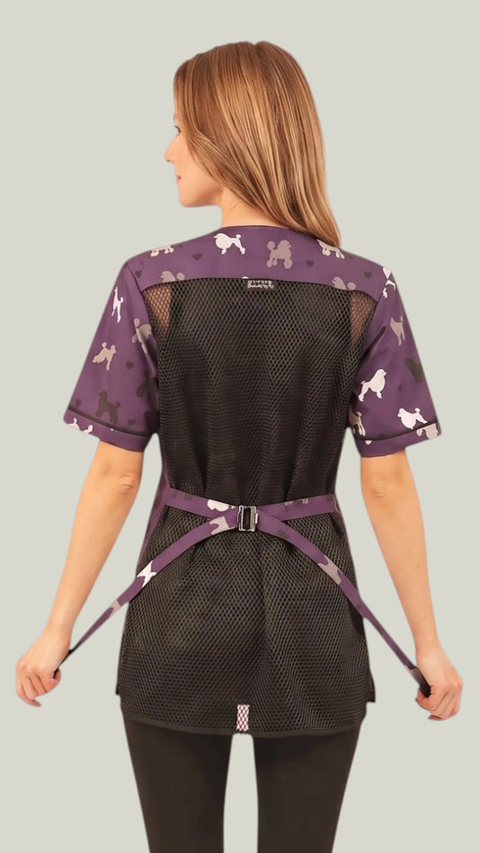 Back view of a light-brown-haired woman holding the back tie belt while modeling the Ladybird Line 710 Purple Poodle Design Waterproof & Bleach Resistant Mesh Back Grooming Smock, showing the black mesh back panel, purple poodle-print shoulder yoke, purple poodle-print short sleeves, and purple poodle-print back tie belt, worn over black pants against a light grey background.