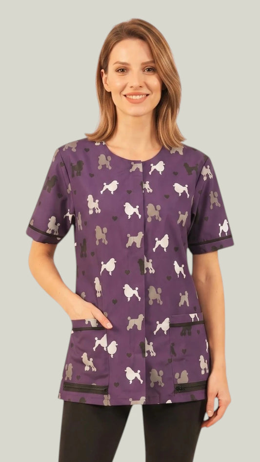 Front view of a smiling light-brown-haired woman modeling the Ladybird Line 710 Purple Poodle Design Waterproof & Bleach Resistant Mesh Back Grooming Smock, featuring a purple short-sleeve grooming smock with a colorful poodle silhouette print, covered front zipper, and deep lapelled front pockets with black trim, worn over black pants against a light grey background.