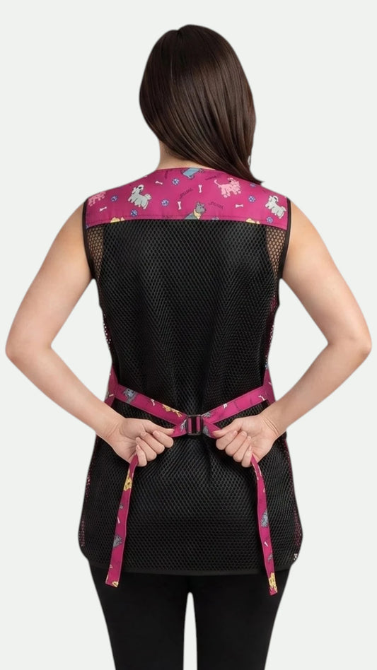 Back view of a woman with dark hair typing her back tie belt while modeling the Ladybird Line 711 Pink Dog Design Waterproof & Bleach Resistant Mesh Back Grooming Vest, showing the black mesh back panel, pink dog-print shoulder yoke, and pink dog-print back tie belt, worn over black pants against a light grey background.