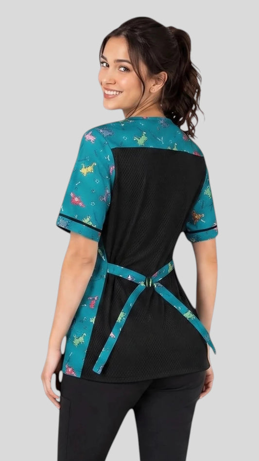 Back view of a woman with dark hair in a bun looking over her shoulder while modeling the Ladybird Line 713 Blue Dog Design Waterproof & Bleach Resistant Mesh Back Grooming Smock, showing the black mesh back panel, teal dog-print shoulder yoke, short sleeves, and back tie belt, worn over black pants against a light grey background.