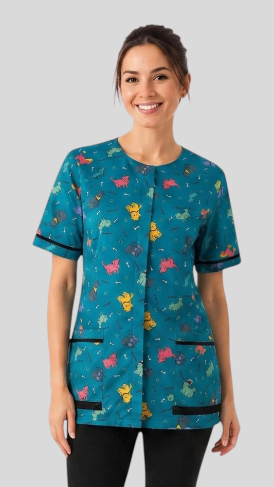 Front view of a smiling woman with dark hair in a bun modeling the Ladybird Line 713 Blue Dog Design Waterproof & Bleach Resistant Mesh Back Grooming Smock, featuring a teal blue short-sleeve grooming smock with a colorful dog silhouette print, covered front zipper, and deep front lapelled pockets with black trim, worn over black pants against a light grey background.