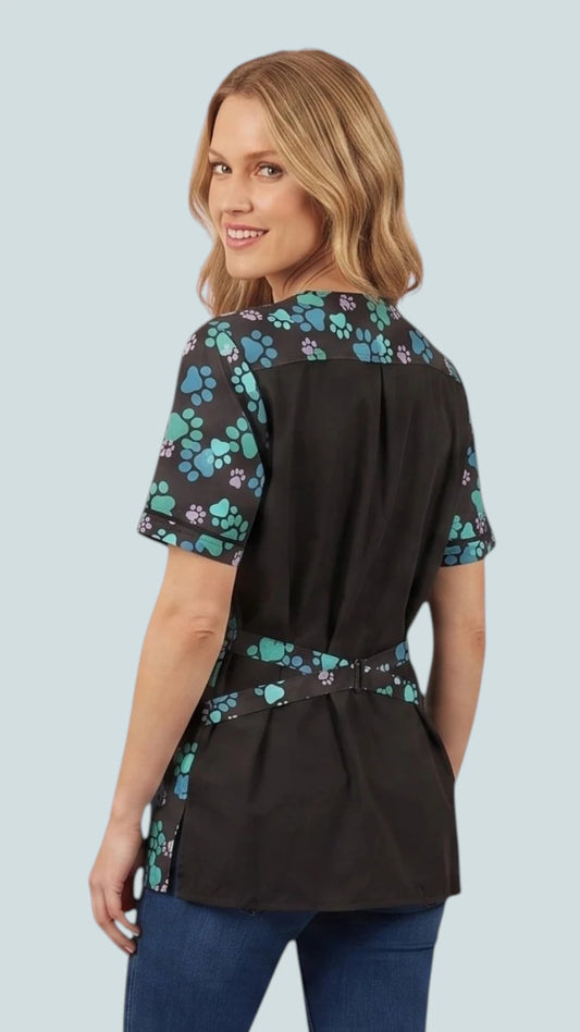 Back view of a blonde woman looking over her shoulder while modeling the Ladybird Line 714 Blue Paw Print Waterproof & Bleach Resistant Solid Back Grooming Smock, showing the blue, teal, and purple paw print pattern on the upper shoulder yoke, sleeves, and back tie belt against a solid black back panel, worn over blue jeans against a light grey background.