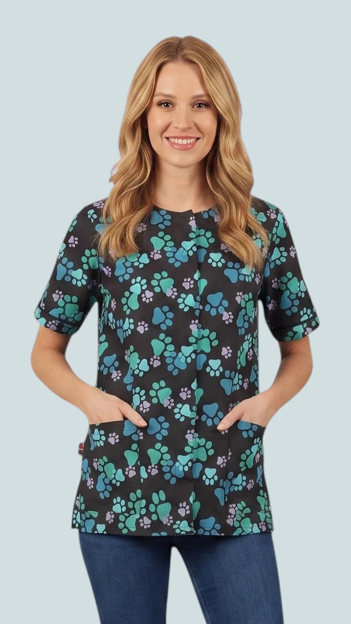 Front view of a smiling blonde woman posing with hands in pockets modeling the Ladybird Line 714 Blue Paw Print Waterproof & Bleach Resistant Solid Back Grooming Smock, featuring a black short-sleeve grooming smock with a blue, teal, and purple paw print pattern, covered front zipper, and deep front pockets, worn over blue jeans against a light grey background.