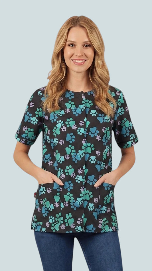 Front view of a smiling blonde woman posing with hands in pockets modeling the Ladybird Line 714 Blue Paw Print Waterproof & Bleach Resistant Solid Back Grooming Smock, featuring a black short-sleeve grooming smock with a blue, teal, and purple paw print pattern, covered front zipper, and deep front pockets, worn over blue jeans against a light grey background.