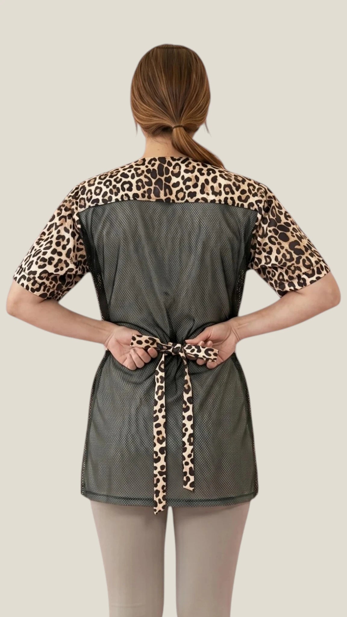 Side profile view of a brown-haired woman posing with hand on hip modeling the Ladybird Line 716 Leopard Print Waterproof & Bleach Resistant Mesh Back Grooming Smock, showing the black side accent panel and leopard print pattern, worn over light-colored pants against a solid light beige background.