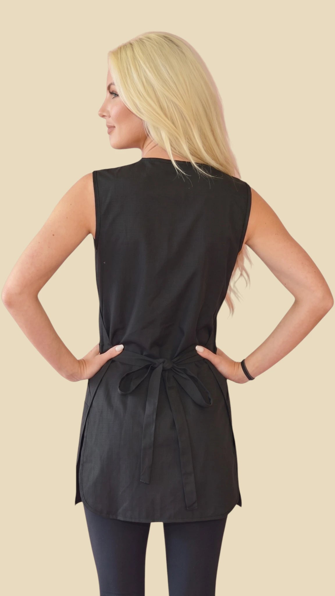 Back view of a blonde woman with hands on hips modeling the Ladybird Line 720 Rhinestone Zipper Water Resistant & Bleach Resistant Cobbler Apron, showing the back waist tie belt closure, worn over black leggings against a solid light beige background.
