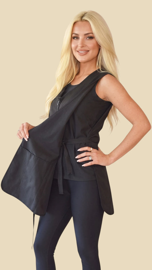 Side view of a blonde woman holding open the front panel of the Ladybird Line 720 Rhinestone Zipper Water Resistant & Bleach Resistant Cobbler Apron, showing the inner tie belt closure, rhinestone zipper accent, and deep front pockets, worn over black leggings against a solid light beige background.