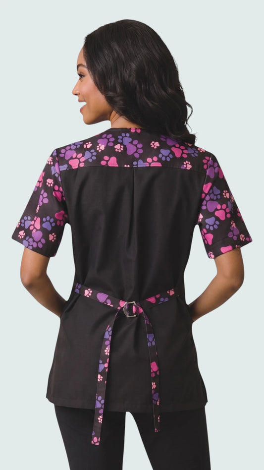 Back view of a woman with dark hair looking over her shoulder while modeling the Ladybird Line 725 Pink & Purple Paw Print Waterproof & Bleach Resistant Solid Back Jacket, showing the pink and purple paw print pattern on the upper yoke, sleeves, and back tie belt against a solid black back panel, worn over black pants against a light grey background.