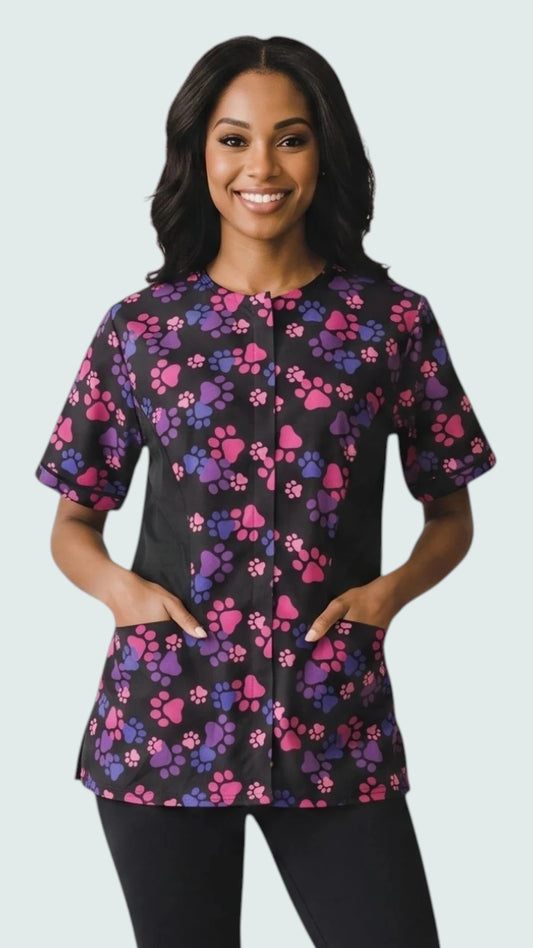 Front view of a smiling woman with dark hair modeling the Ladybird Line 725 Pink & Purple Paw Print Waterproof & Bleach Resistant Solid Back Jacket, featuring a black short-sleeve grooming jacket with a pink and purple paw print pattern, covered front zipper, and deep pockets, worn over black pants against a light grey background.