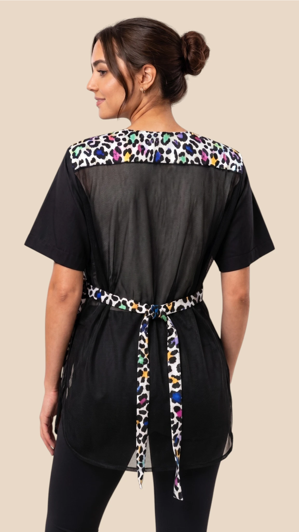Back view of a dark-haired woman modeling the Ladybird Line 737 Multi Color Leopard Color Block Waterproof Grooming Smock, featuring a mesh back panel, multi-color leopard print upper accent and tie belt, worn over black pants against a solid beige background.