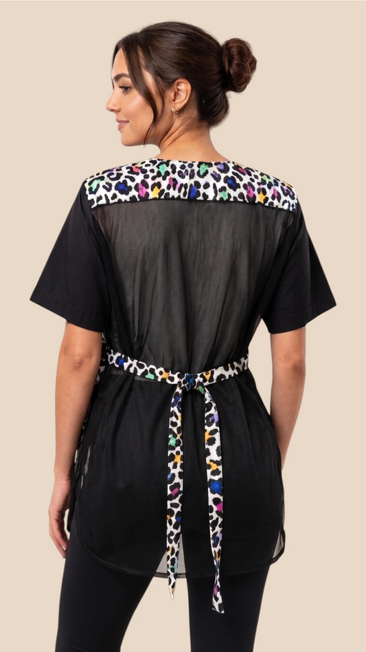 Back view of a dark-haired woman modeling the Ladybird Line 737 Multi Color Leopard Color Block Waterproof Grooming Smock, featuring a mesh back panel, multi-color leopard print upper accent and tie belt, worn over black pants against a solid beige background.