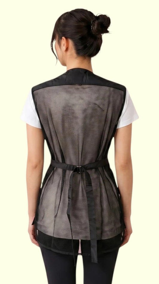 Back view of a dark-haired woman in a hair bun modeling the Ladybird Line 745 Ladies Black Waterproof Hair Resistant & Bleach Resistant Mesh Back Vest, showing the full black mesh back panel and adjustable back belt strap with buckle closure, worn over a white t-shirt and black pants against a light beige background.