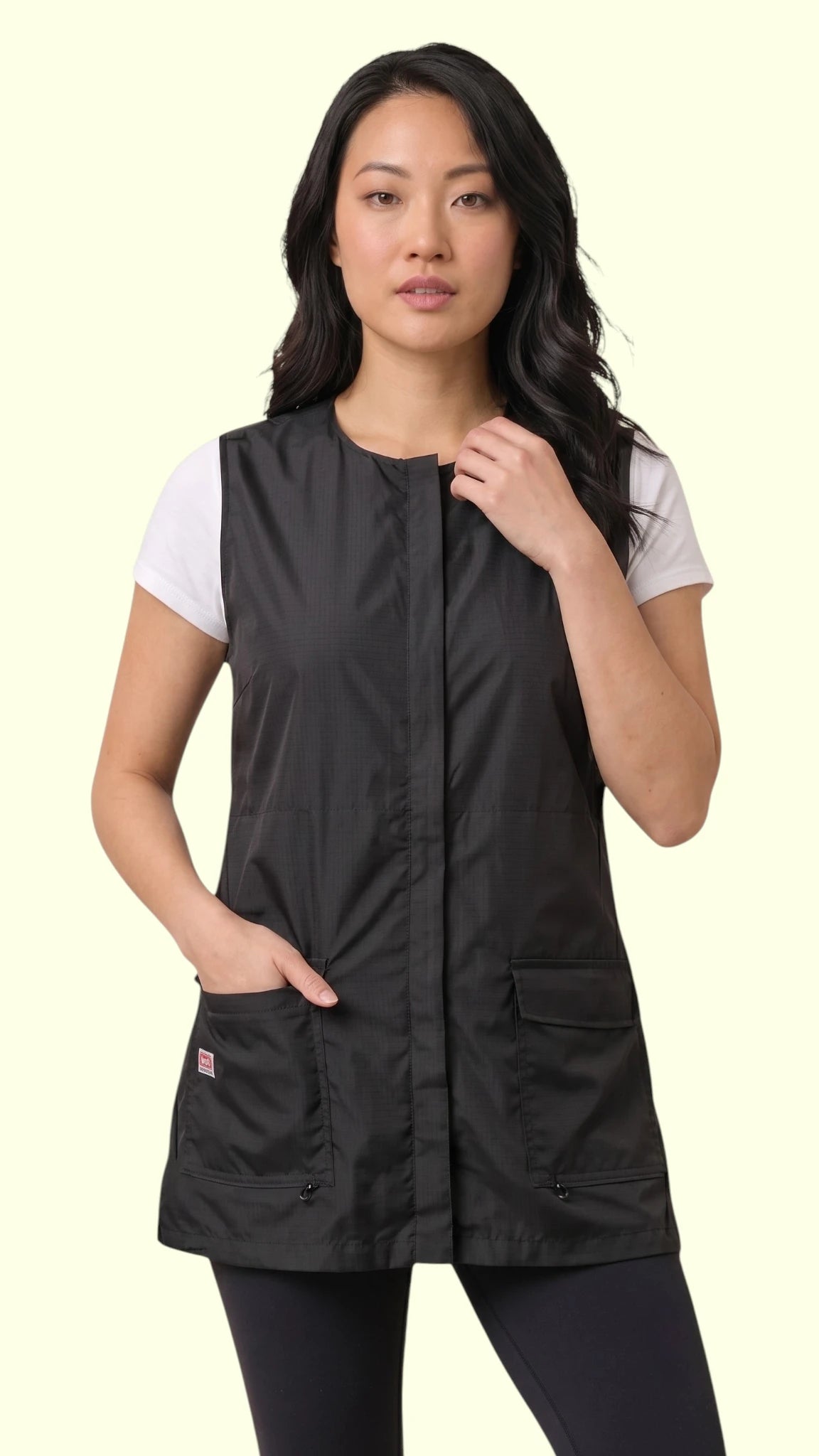 Front view of a dark-haired woman modeling the Ladybird Line 745 Ladies Black Waterproof Hair Resistant & Bleach Resistant Mesh Back Vest, featuring a sleeveless black grooming vest with covered front zipper and deep flap pockets, worn over a white t-shirt and black pants against a light beige background.