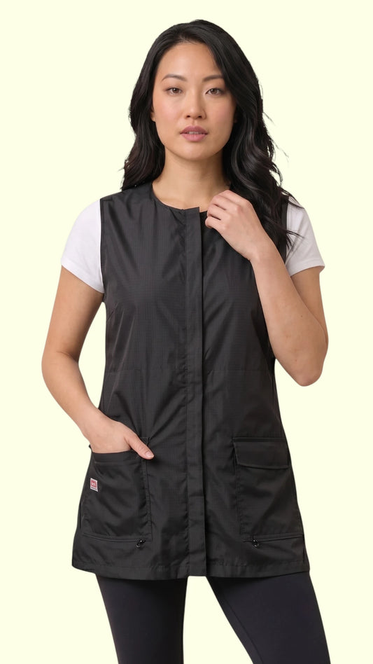 Front view of a dark-haired woman modeling the Ladybird Line 745 Ladies Black Waterproof Hair Resistant & Bleach Resistant Mesh Back Vest, featuring a sleeveless black grooming vest with covered front zipper and deep flap pockets, worn over a white t-shirt and black pants against a light beige background.