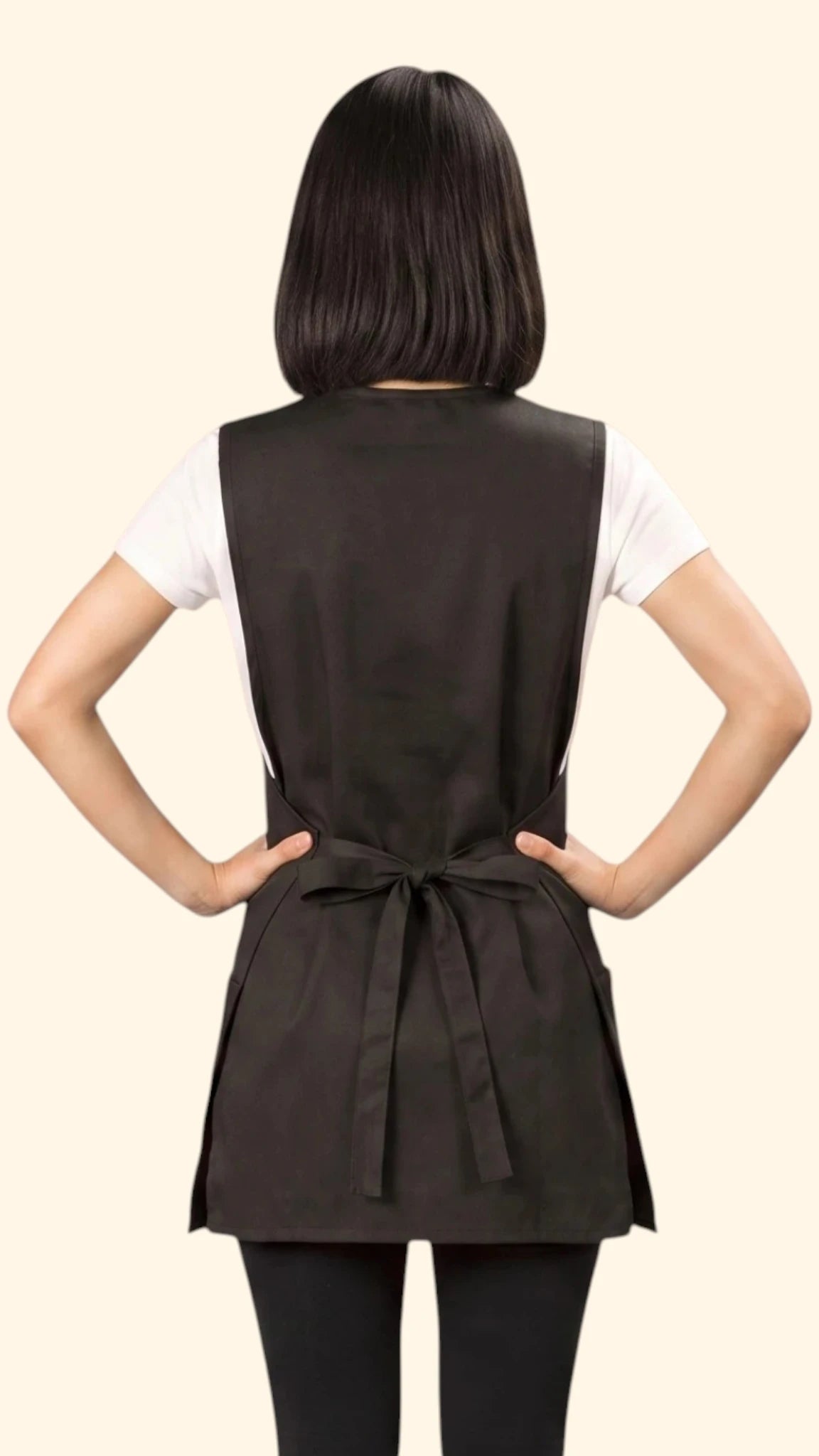 Back view of a dark-haired woman with hands on hips modeling the Ladybird Line 750 Pro Apron Water Resistant & Bleach Resistant Cobbler Style Apron, showing the back waist tie belt closure, worn over a white t-shirt and black pants against a solid light beige background.
