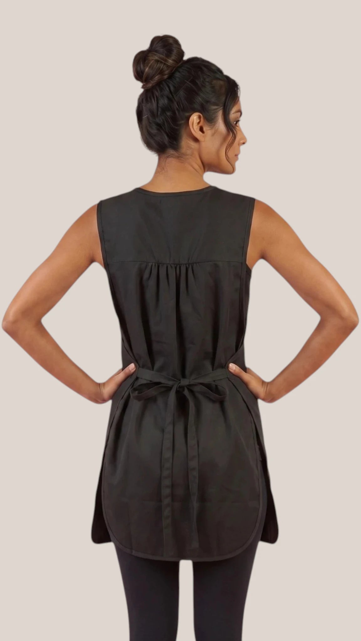 Back view of a dark-haired woman in a hair bun with hands on hips modeling the Ladybird Line 775 Waterproof Hair Resistant & Bleach Resistant Regular Size Cobbler Apron, showing the back waist tie belt closure, worn over black leggings against a light beige background.