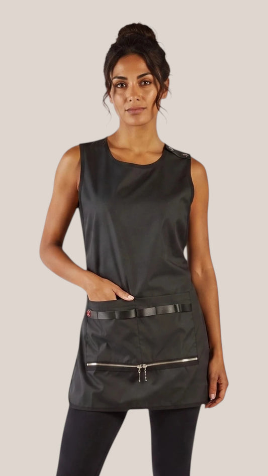 Front view of a dark-haired woman in a hair bun modeling the Ladybird Line 775 Waterproof Hair Resistant & Bleach Resistant Regular Size Cobbler Apron, featuring a sleeveless black cobbler apron with deep front pockets and zippered bottom closures, worn over black leggings against a light beige background.