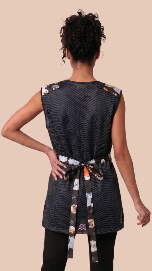 Back view of a dark-haired woman in a hair bun modeling the Ladybird Line 777 Dog Groups Waterproof & Bleach Resistant Grooming Vest with Mesh Back, showing the black mesh back panel, printed upper shoulder yoke, and printed back tie belt, worn over dark pants against a solid light beige background.