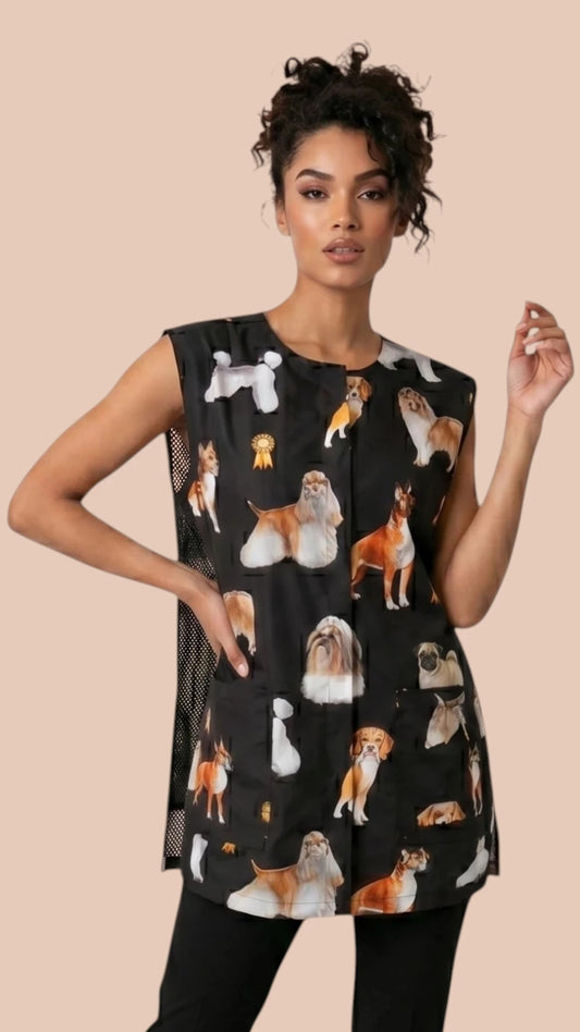 Front view of a dark-haired woman in a hair bun modeling the Ladybird Line 777 Dog Groups Waterproof & Bleach Resistant Grooming Vest with Mesh Back, featuring a sleeveless black grooming vest with an all-over print of various dog breed groups, worn over black pants against a solid light beige background.