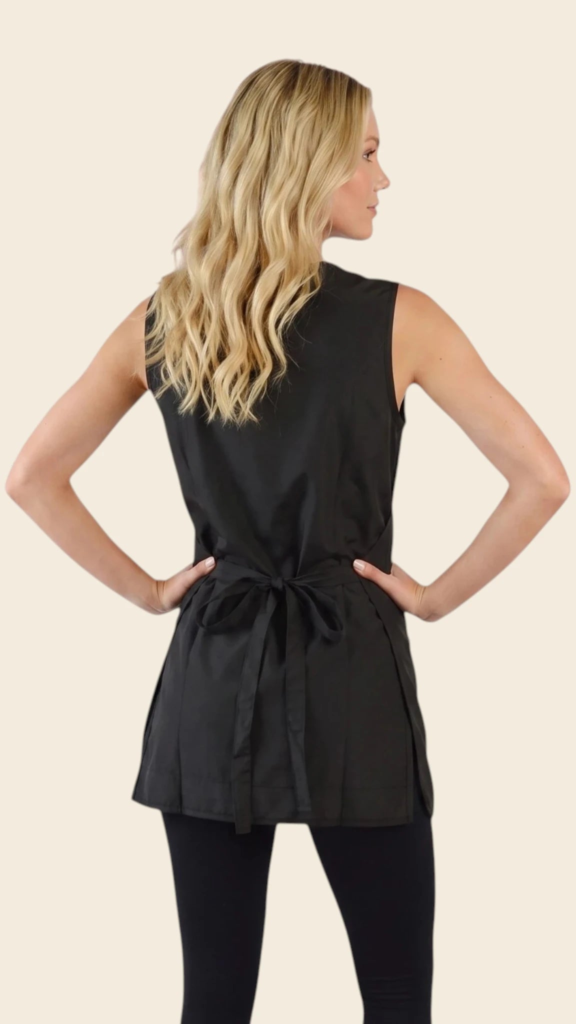 Back view of a blonde woman with hands on hips modeling the Ladybird Line 800 Waterproof Hair Resistant & Bleach Resistant Curvy Size Cobbler Apron, showing the back tie belt cinched at the waist, worn over black leggings against a solid light beige background.