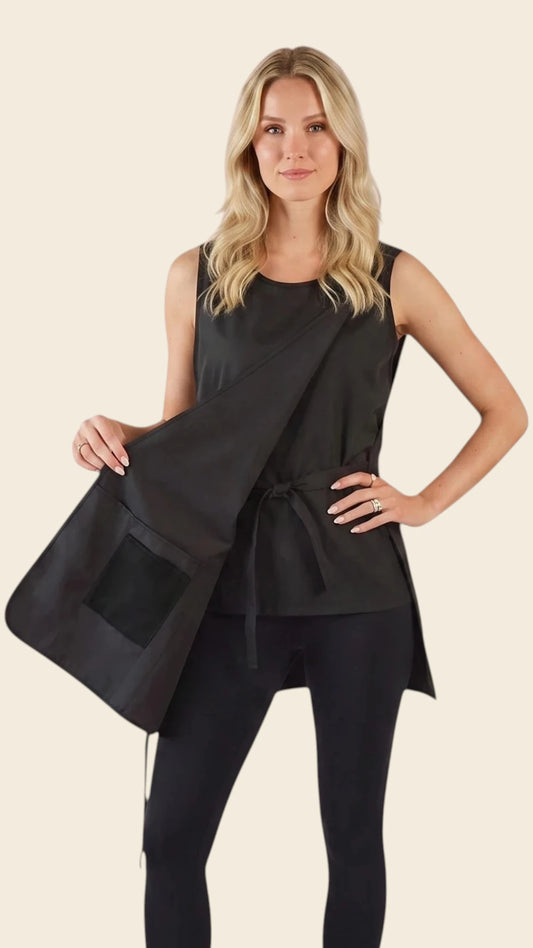 Side view of a blonde woman holding open the side wrap panel of the Ladybird Line 800 Waterproof Hair Resistant & Bleach Resistant Curvy Size Cobbler Apron, showing the inner tie closure and deep front pocket, worn over black leggings against a light beige background.