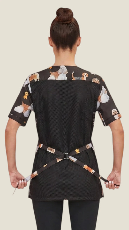 Back view of a dark-haired woman in a hair bun modeling the Ladybird Line 803 Dog Groups Water Resistant & Bleach Resistant Stylist Jacket with Mesh Back, showing the black mesh back panel, printed upper shoulder yoke, short sleeves, and printed back tie belt, worn over dark pants against a light beige background.