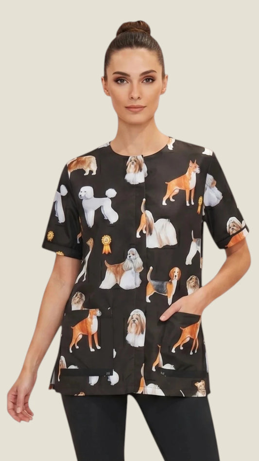 Front view of a dark-haired woman in a hair bun modeling the Ladybird Line 803 Dog Groups Water Resistant & Bleach Resistant Stylist Jacket with Mesh Back, featuring a black short-sleeve grooming jacket with an all-over print of various dog breed groups, worn with black pants against a solid light beige background.