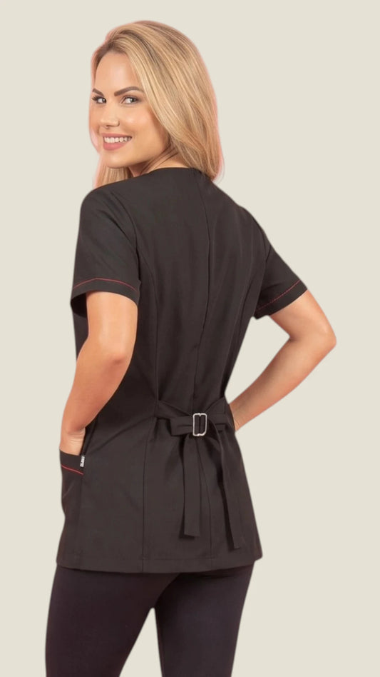 Back view of a smiling blonde woman looking back over her shoulder while modeling the Ladybird Line 825 Black Water Resistant & Bleach Resistant Styling Jacket with Red Zipper, showing the adjustable back belt strap with buckle closure and short sleeves with red contrast stitching, worn over black leggings against a light beige background.