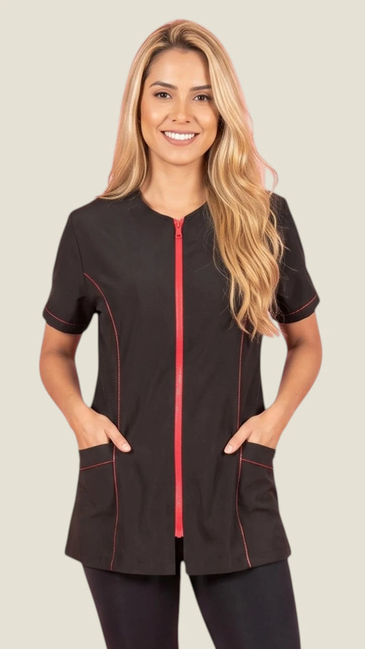 Front view of a smiling blonde woman modeling the Ladybird Line 825 Black Water Resistant & Bleach Resistant Styling Jacket with Red Zipper, featuring a short-sleeve black grooming jacket with vibrant red contrast stitching, red front zipper, and deep front pockets, worn against a light beige background.