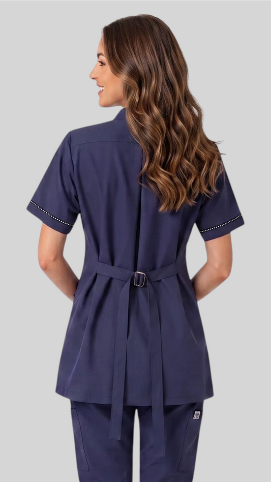 Back view of a brunette woman modeling the Ladybird Line 854 Navy Rhinestone Trim Waterproof Grooming Smock, showing the adjustable tie belt at the waist and short sleeves over matching navy pants against a neutral grey background.