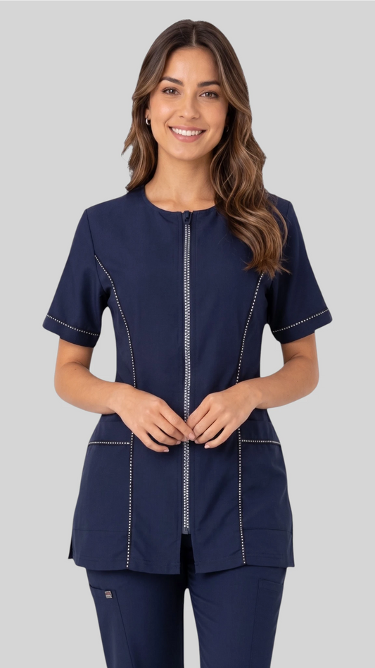 Front view of a brunette woman smiling while modeling the Ladybird Line 854 Navy Rhinestone Trim Waterproof Grooming Smock, featuring a navy short-sleeve zip-up smock with rhinestone trim along the zipper, sleeves, and pockets, paired with matching navy pants against a neutral grey background.