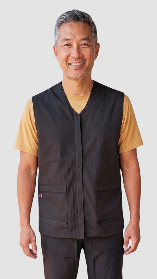 Front view of an Asian man with short grey hair smiling while modeling the Ladybird Line 880 Black V-Neck Waterproof & Bleach Resistant Mesh Back Vest, featuring a sleeveless black grooming vest with front flap pockets and zippered bottom closures, worn over a gold t-shirt against a light grey background.