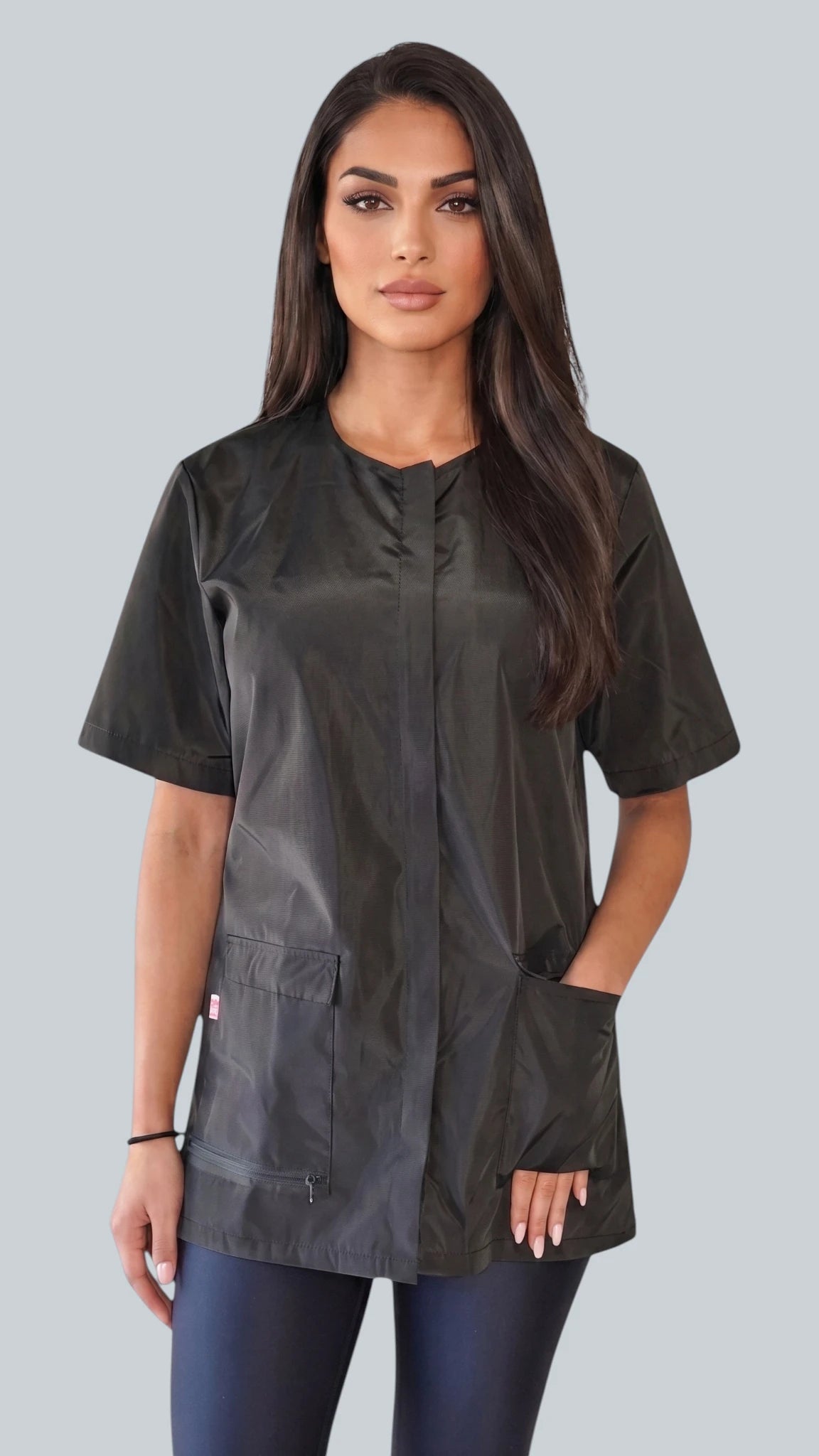 Front view of a dark-haired woman posing with hands in pockets modeling the Ladybird Line 950 Classic Black Waterproof & Bleach Resistant Grooming Smock, featuring a short-sleeve black grooming jacket with covered front zipper and front pockets, worn over dark blue pants against a light grey background.